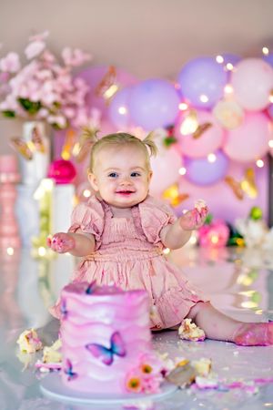 Smiling baby girl in pink dress celebrates first birthday with cake smash, surrounded by balloons and butterfly decorations.
