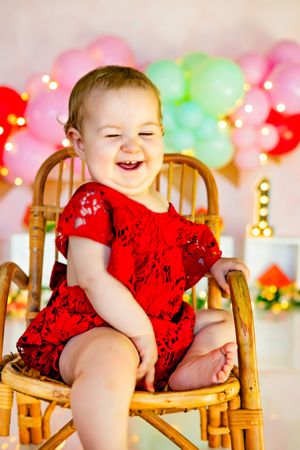 A joyful baby girl in a red lace dress laughs while sitting in a rattan chair surrounded by colorful balloons.