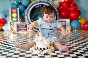 Baby boy smashing first birthday cake on checkered floor with colorful balloon backdrop.