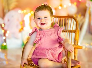 Smiling baby girl in pink ruffle romper sitting in a rattan chair with bokeh lights in background.