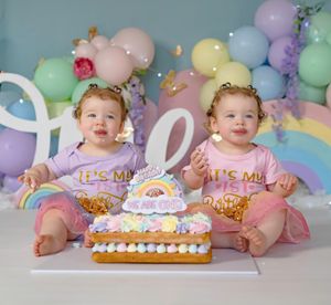 Twin baby girls in matching pink outfits celebrate their first birthday with a rainbow cake and pastel balloon backdrop.