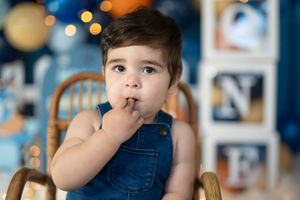 Toddler boy in denim overalls sitting in wicker chair at a blue and gold first birthday party celebration.