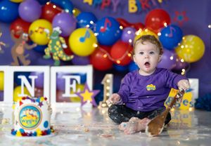 Baby in purple Wiggles outfit sits among smashed cake, colorful balloons, and letter blocks at a Wiggles-themed birthday party.