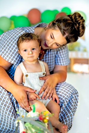 A smiling mother holds her baby at a first birthday party with green balloons in the background.