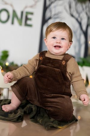 Smiling baby in brown corduroy overalls sits in a festive first birthday photo setup with ONE sign and bokeh lights.