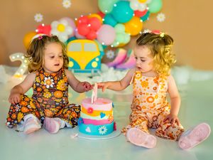 Two toddler girls in floral dresses touching a colorful birthday cake with balloon decorations behind them.