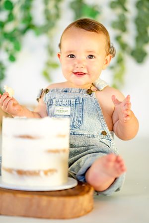 Adorable baby in denim overalls smiling during first birthday cake smash photo session with white cake.