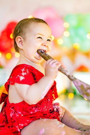 Happy baby girl in red sequin dress eating cake with a spoon at her first birthday party celebration.