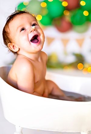 Happy baby laughing in a small white bathtub with colorful Christmas bokeh lights in the background.
