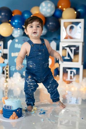 Toddler in denim overalls smashes first birthday cake surrounded by blue and gold balloon decorations.