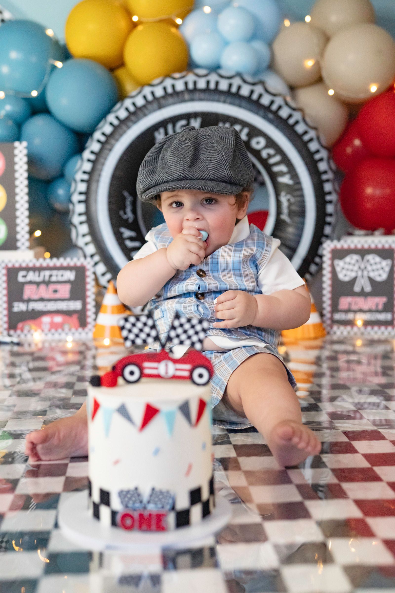 Baby boy in newsboy cap celebrates first birthday with race car themed cake and colorful balloon backdrop.
