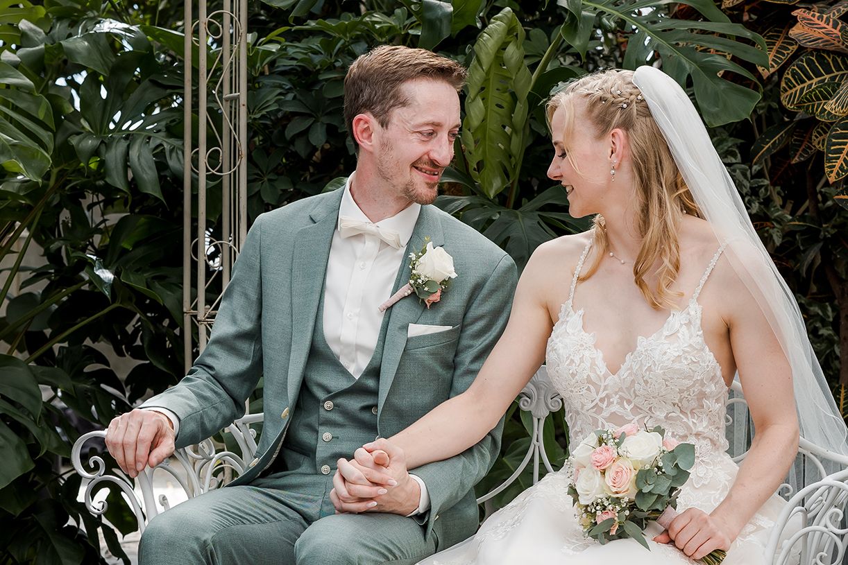 A bride and groom sit together on a white bench, smiling at each other, surrounded by lush greenery.