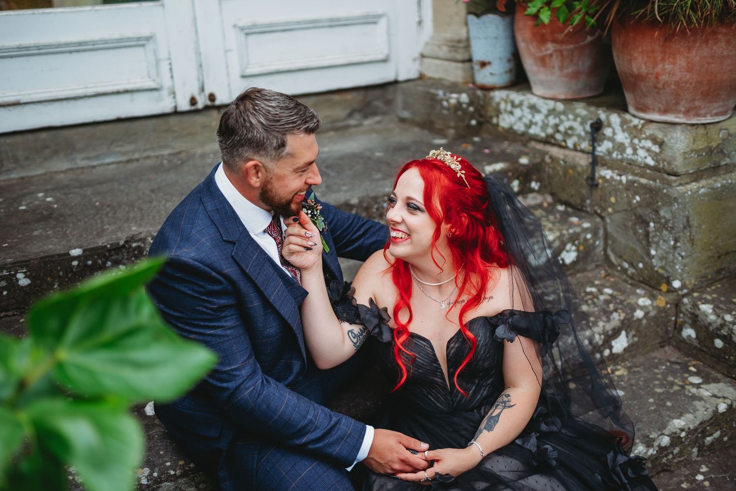 Couple in wedding attire sitting on stone steps, bride in black gown with red hair, groom in navy suit at Clevedon Hall in Sommerset