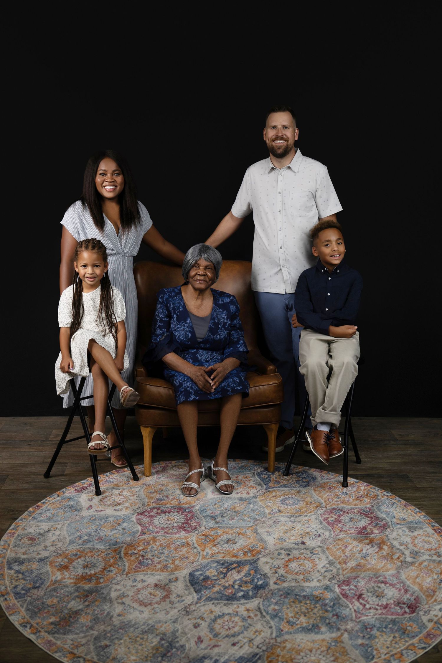 Multi-generational family portrait with five members posing together on a round rug against a dark background.
