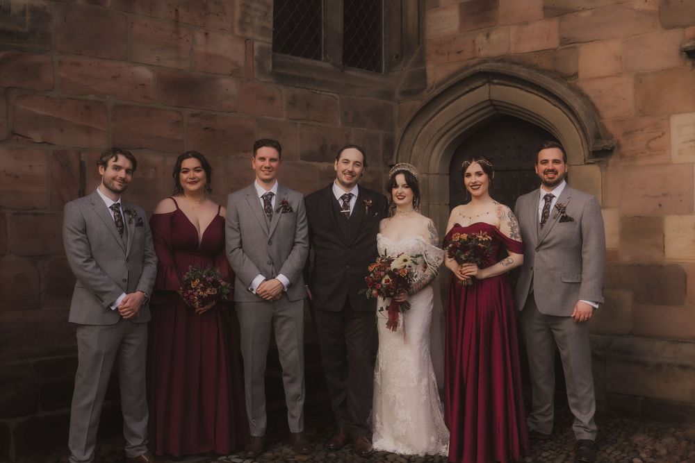 Wedding party poses outside stone church, bride in white lace gown with groomsmen in gray suits and bridesmaids in burgundy dresses.