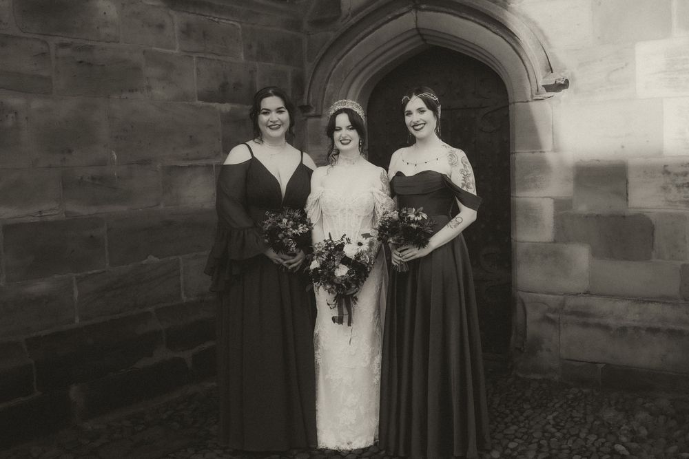 Bride in lace gown with two bridesmaids in dark dresses holding bouquets outside a stone church.