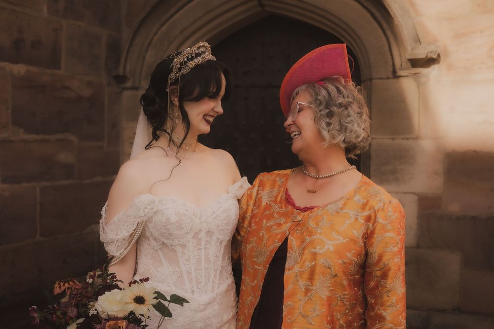 Bride in white lace gown with tiara holds bouquet beside smiling woman in orange dress and pink hat outside stone church.