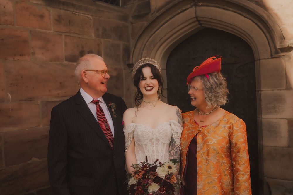 Bride in lace gown and tiara poses with smiling older couple outside stone church door.