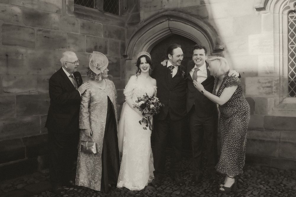 Black and white wedding photo of bride and groom with family outside historic stone church.