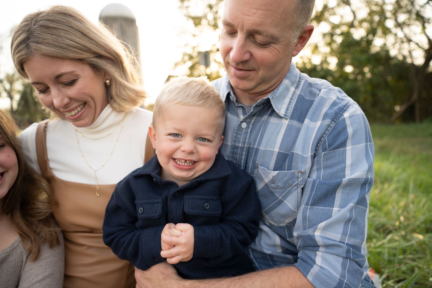 A happy toddler boy smiles while being held by his parents outdoors in a warm family portrait session.