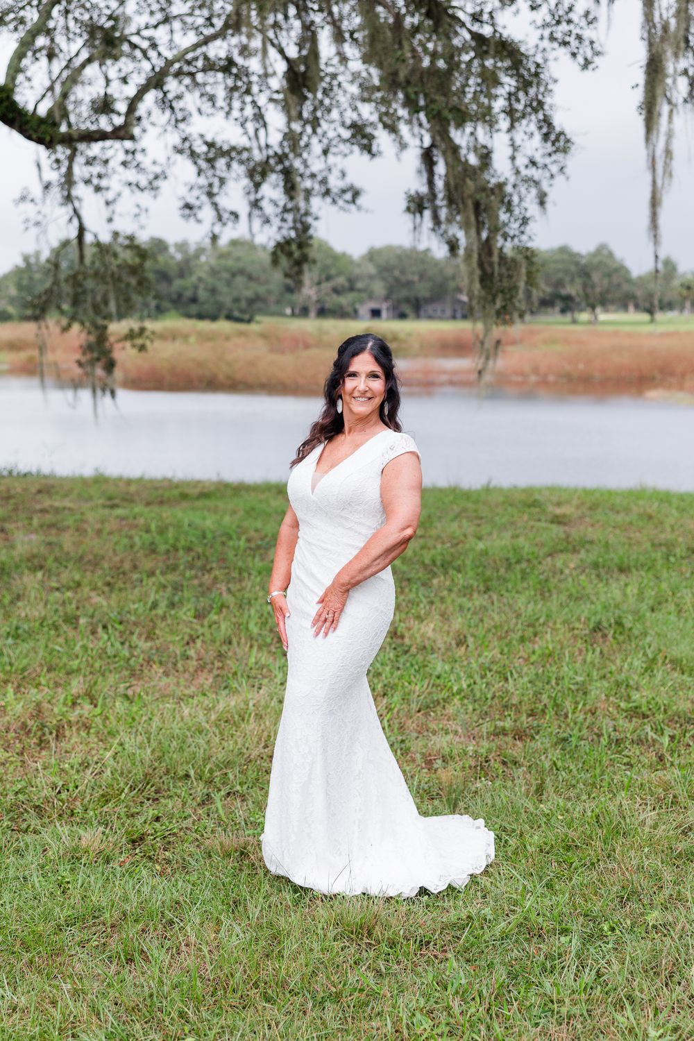 Bride in white lace mermaid gown poses gracefully on green lawn near a serene lake with Spanish moss-draped trees.