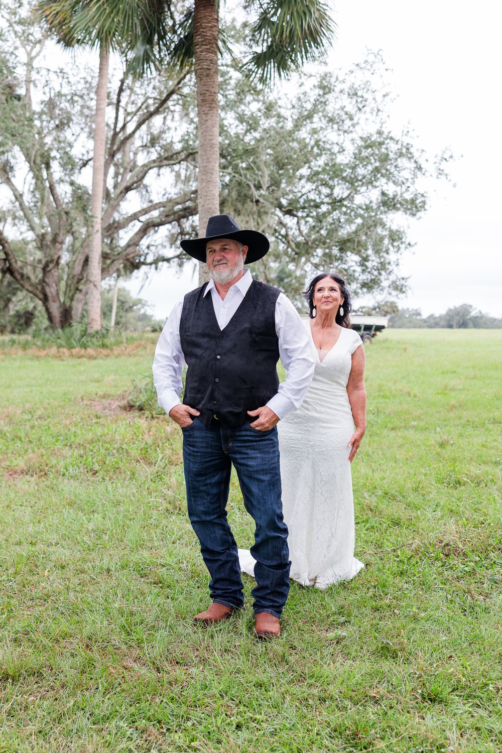 A bride in white dress and groom in cowboy hat and vest pose outdoors on green grass near palm trees.