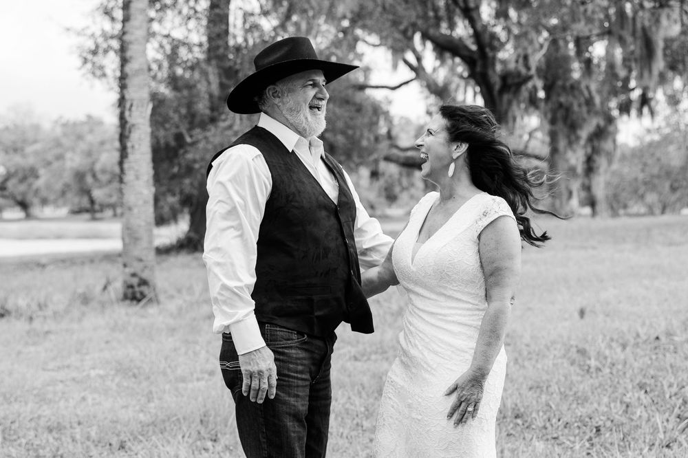Black and white photo of a laughing couple in wedding attire, groom in cowboy hat and vest, bride in white dress, outdoors.