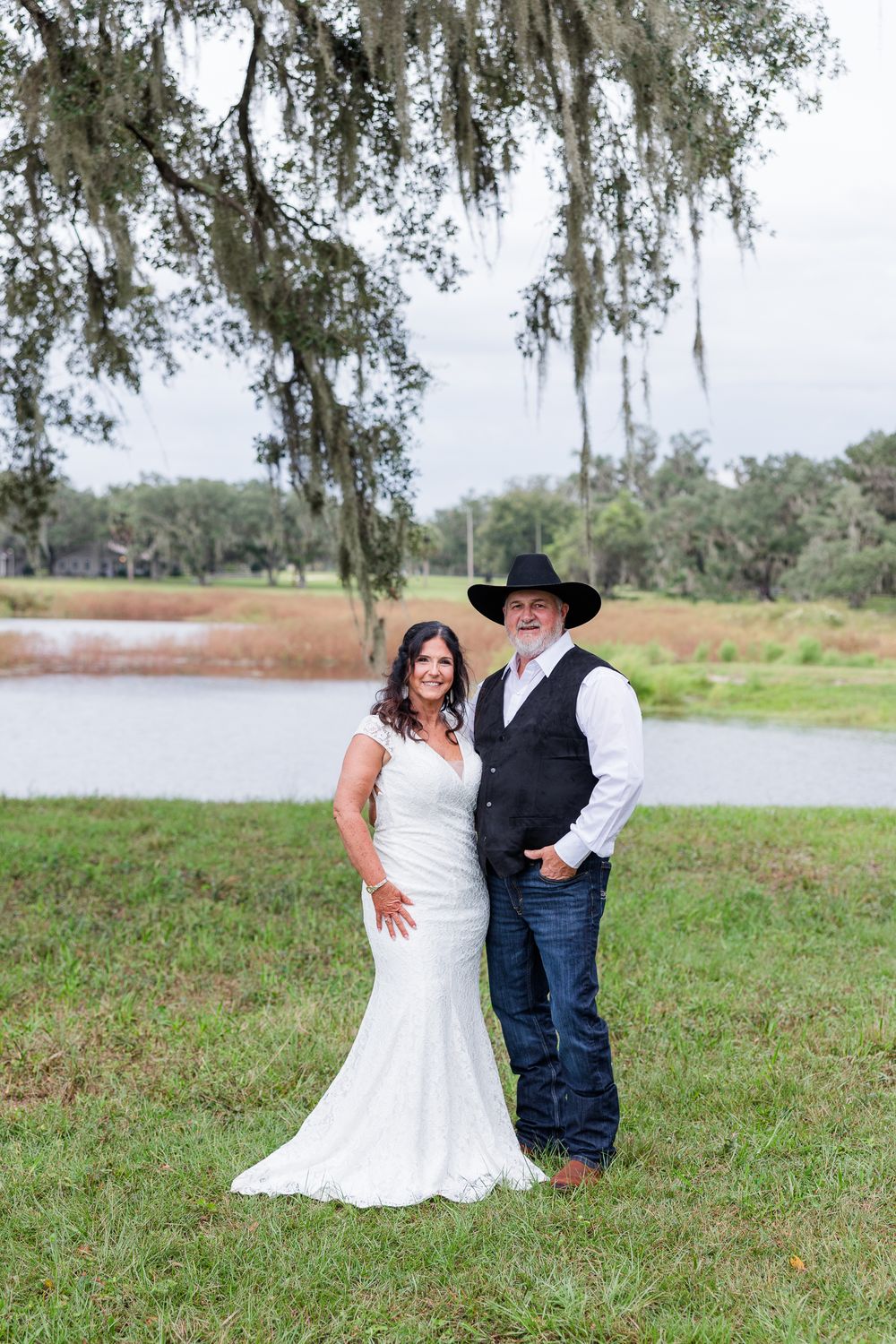 Bride in white mermaid gown and groom in cowboy hat and vest pose by a pond under Spanish moss-draped oak trees.