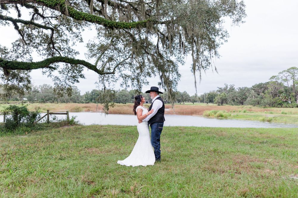 Bride and groom share a romantic embrace under a moss-draped oak tree by a serene pond at an outdoor wedding.