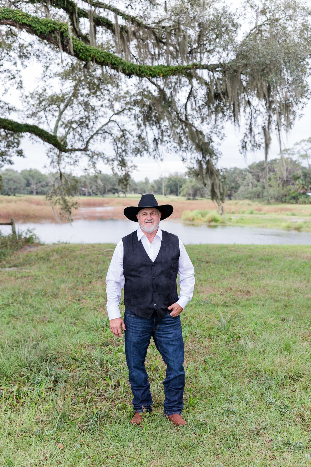 Older man in black cowboy hat, vest, and jeans standing on green grass near a river with Spanish moss trees.