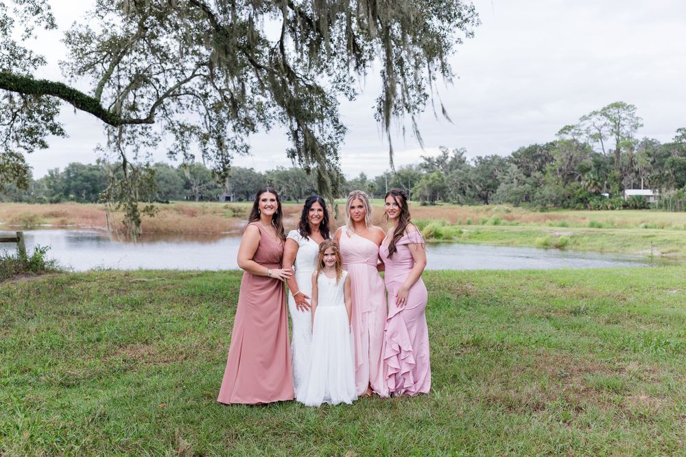 Bride in white gown poses with four bridesmaids in pink dresses outdoors near a pond surrounded by lush greenery.