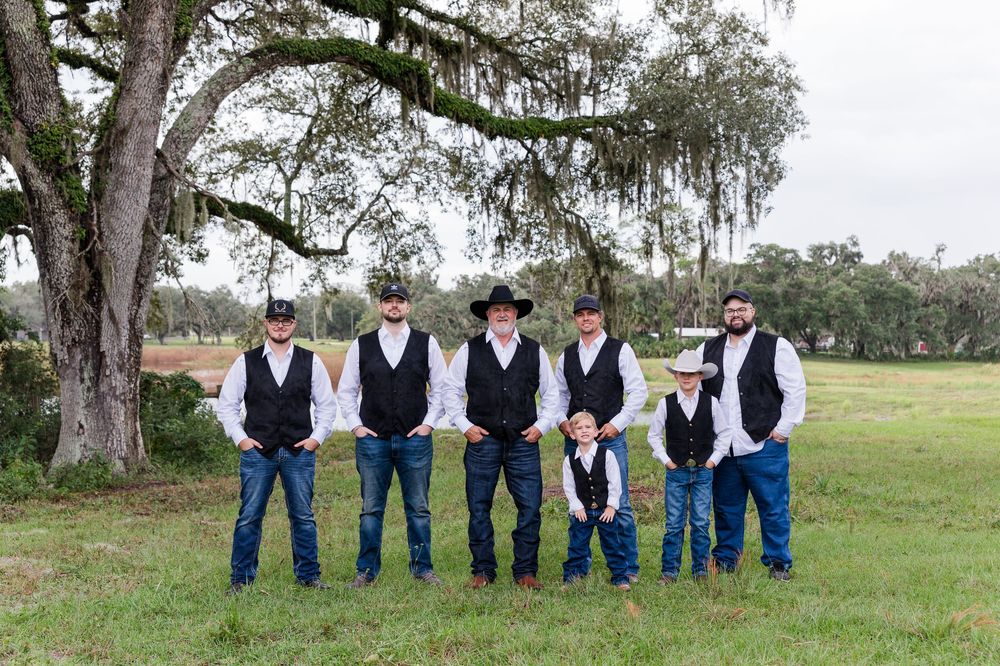 Groomsmen in black vests, jeans, and cowboy hats pose outdoors under a moss-draped oak tree at a country wedding.