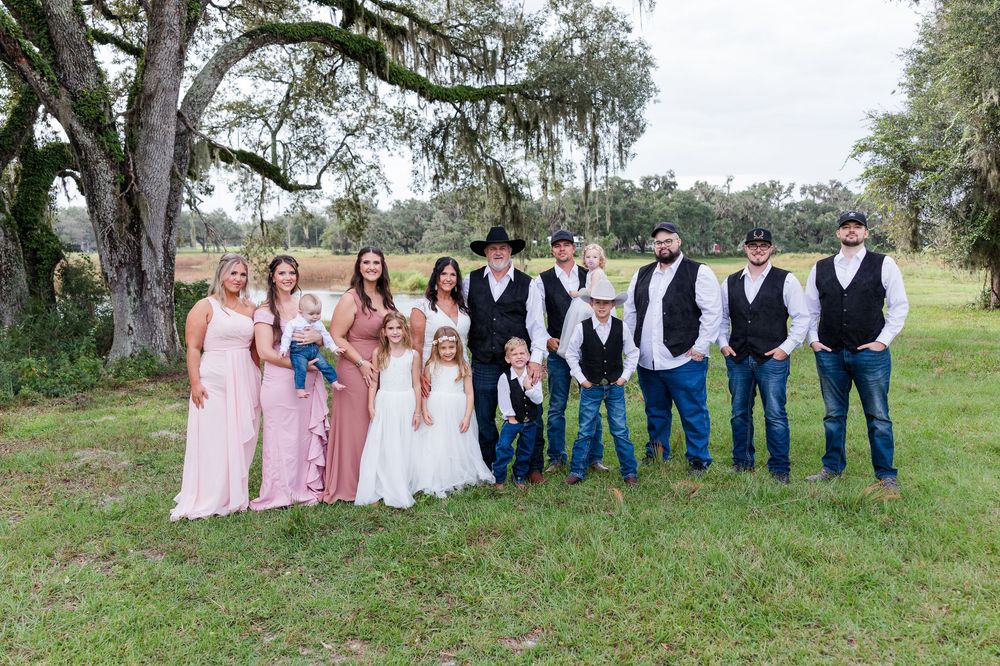 Wedding party poses outdoors under oak trees, bridesmaids in pink dresses, groomsmen in vests and cowboy hats.