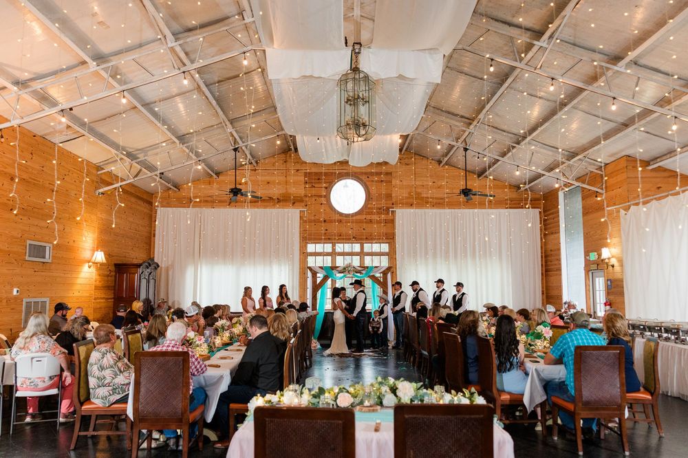 Elegant indoor wedding ceremony in a rustic brick venue with string lights and guests seated at decorated tables.