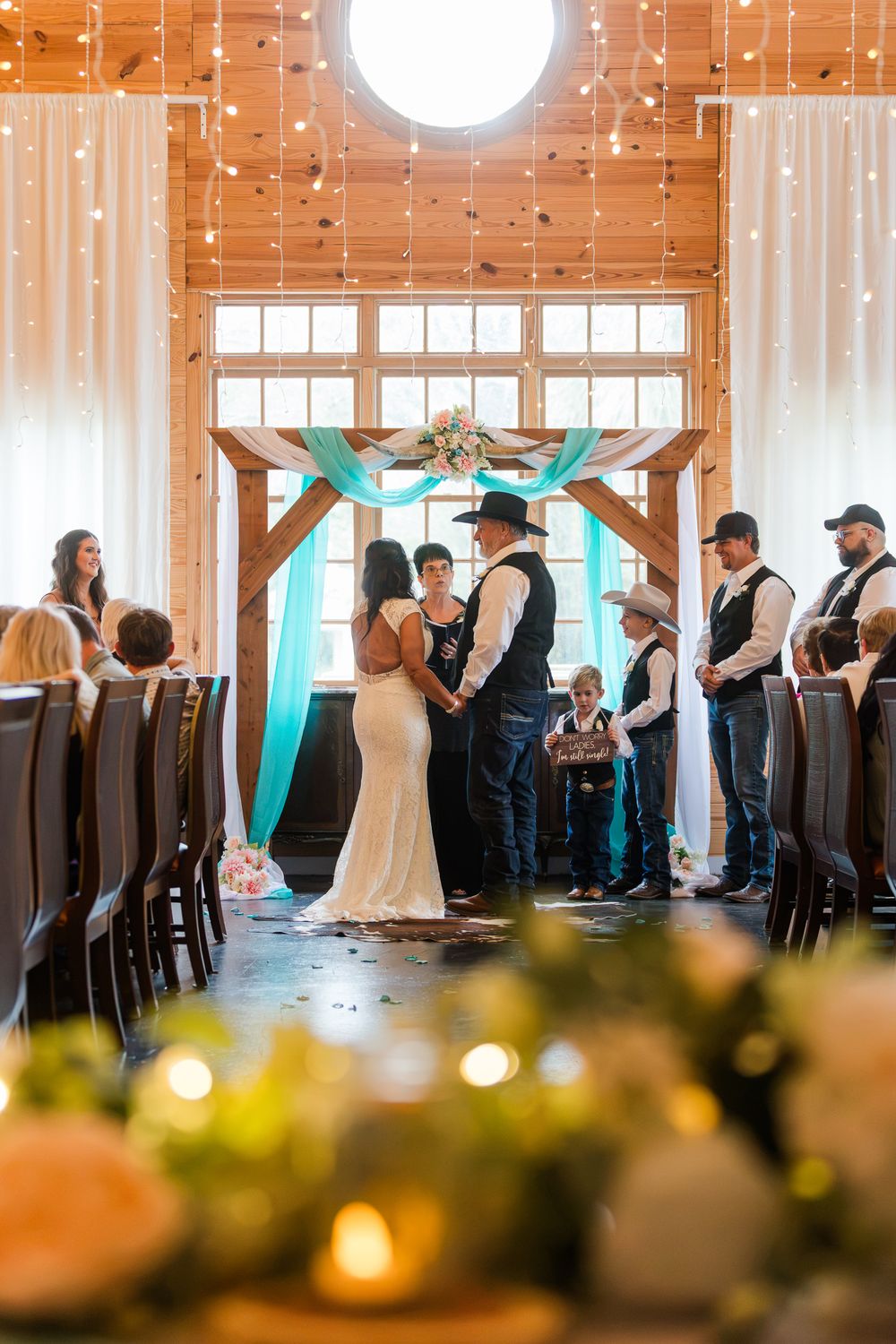 Rustic barn wedding ceremony with couple exchanging vows under a wooden arch with teal draping and string lights.