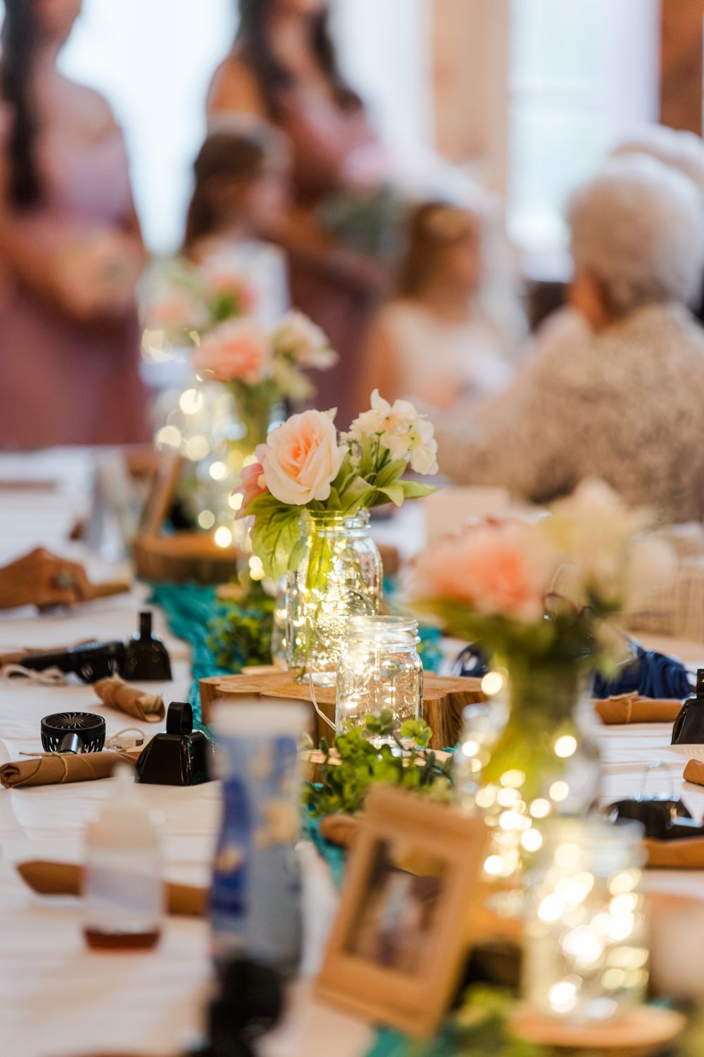 Elegant wedding reception table with pink roses in mason jars, fairy lights, and teal accents on a white tablecloth.