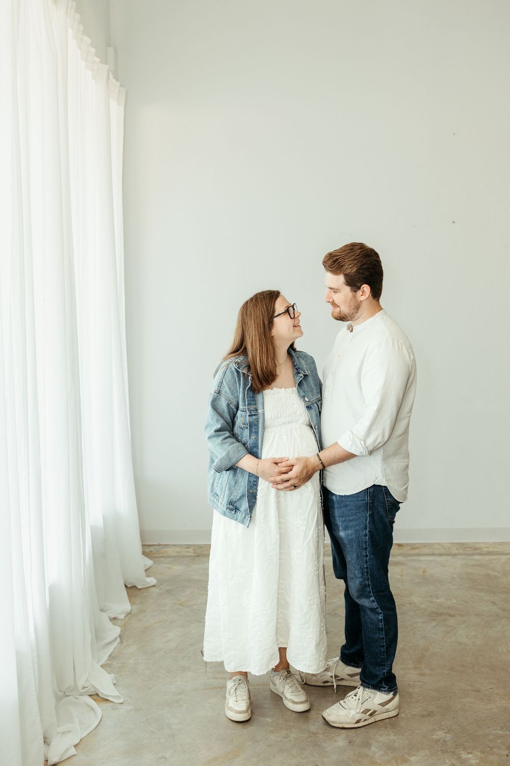 Pregnant couple standing together in a bright white room, man in white shirt and jeans, woman in white dress and denim jacket.