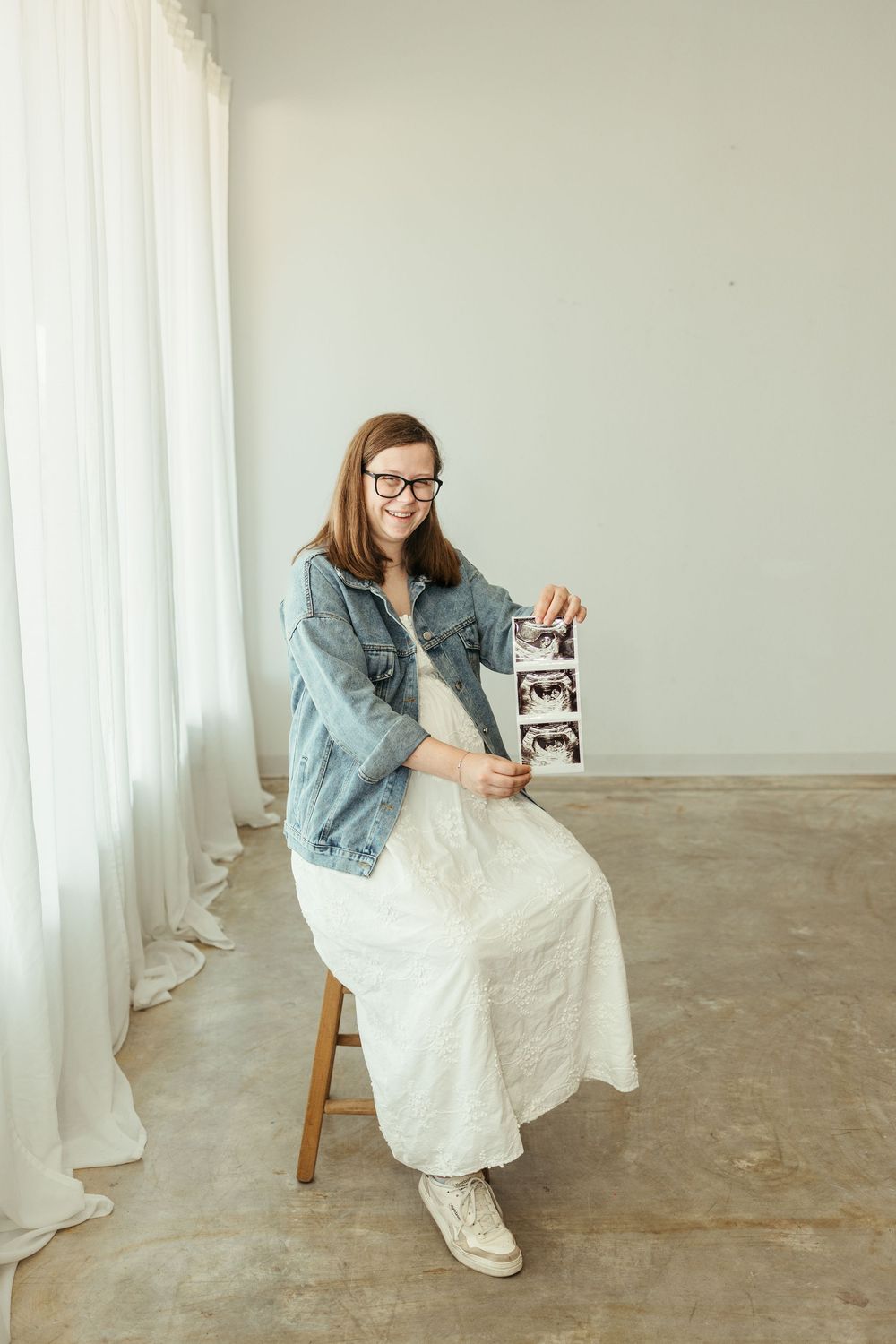 Smiling pregnant woman in denim jacket and white dress sits on stool holding ultrasound photos.