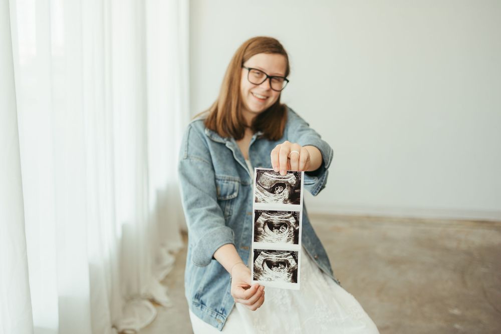 Smiling pregnant woman in glasses holding ultrasound photos, wearing denim jacket, standing near window in bright room.