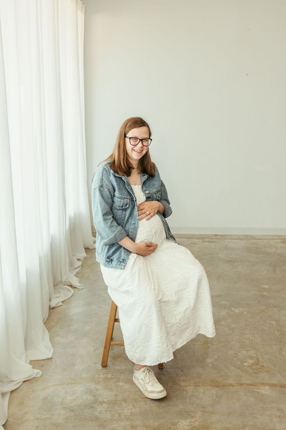Smiling pregnant woman in white dress and denim jacket sits on stool, cradling baby bump in bright studio.