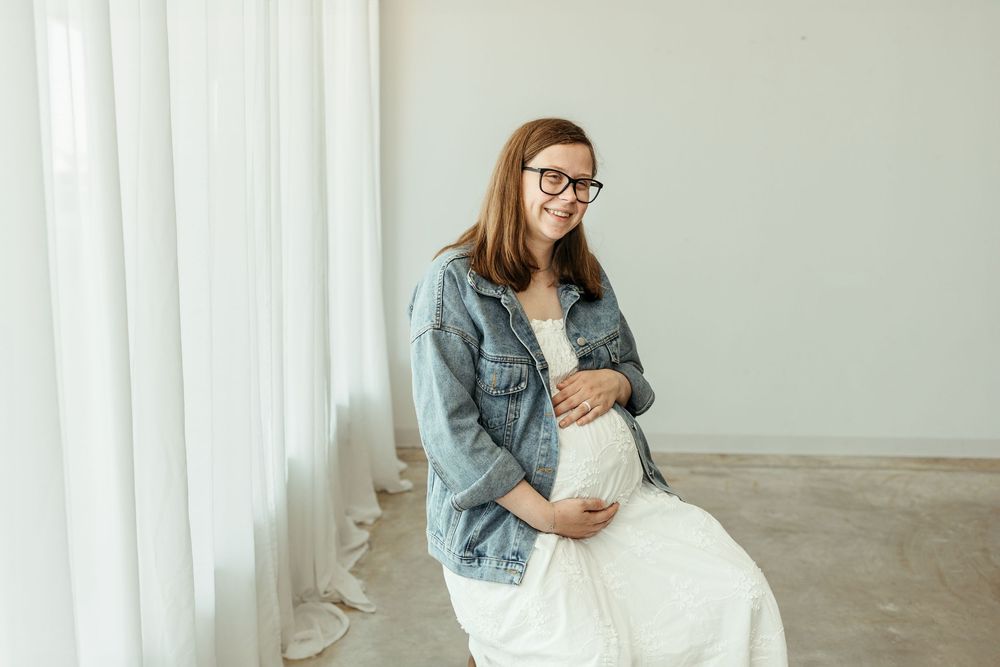 Happy pregnant woman in denim jacket and white dress sitting, cradling baby bump, smiling in bright white room.