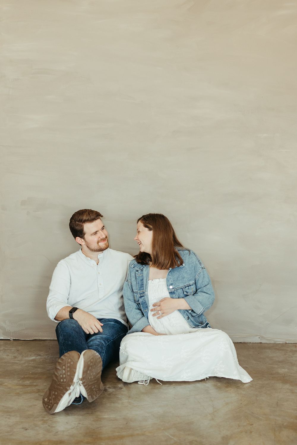 A couple sits on a concrete floor against a gray wall, smiling at each other, the woman visibly pregnant in a white dress.