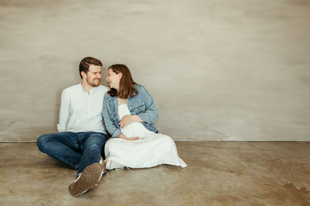Smiling couple sitting on floor, pregnant woman in white dress and denim jacket, man in light blue sweater and jeans.