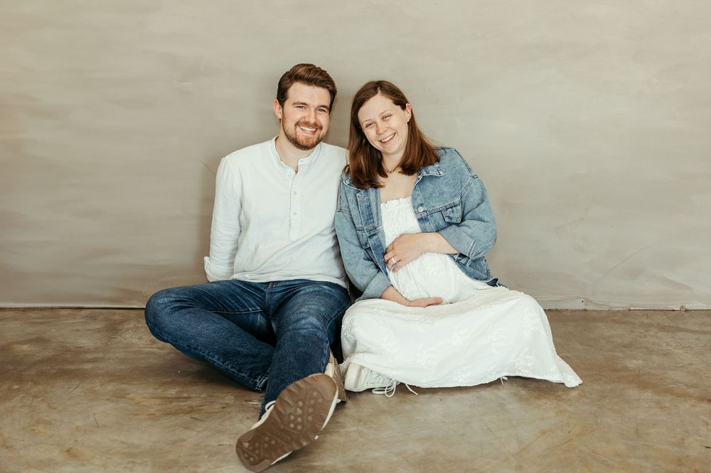 Happy couple sitting on floor, woman holding newborn baby, smiling at camera in a bright studio setting.