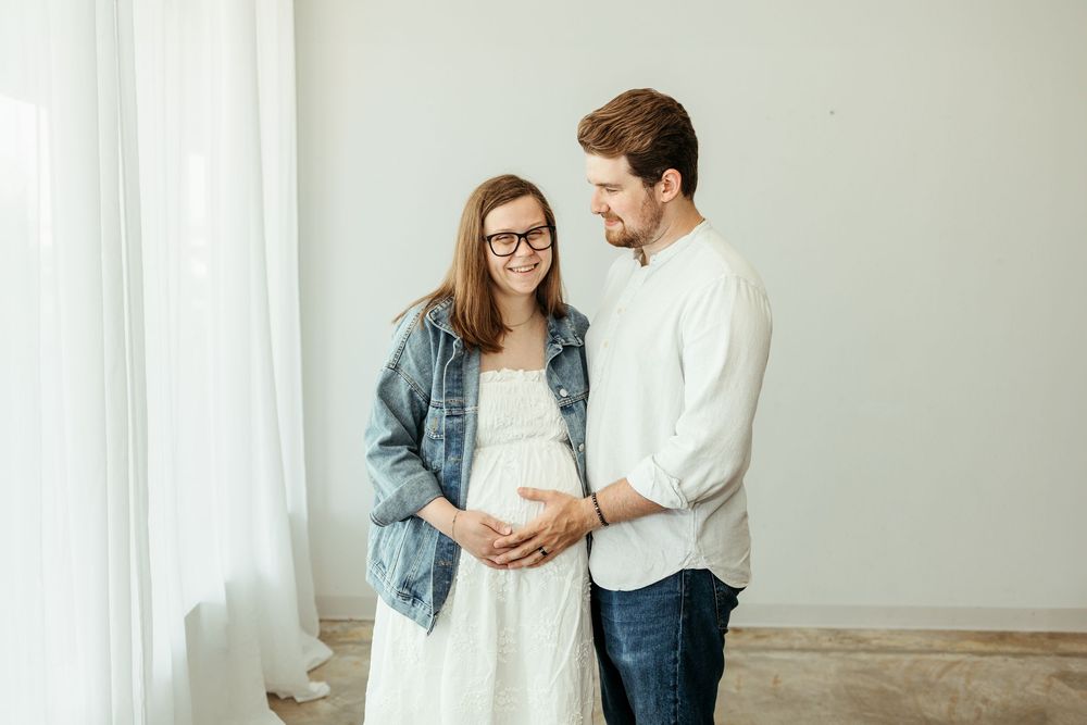 Smiling pregnant couple in white room, man in white shirt touching baby bump, woman in denim jacket and white dress.