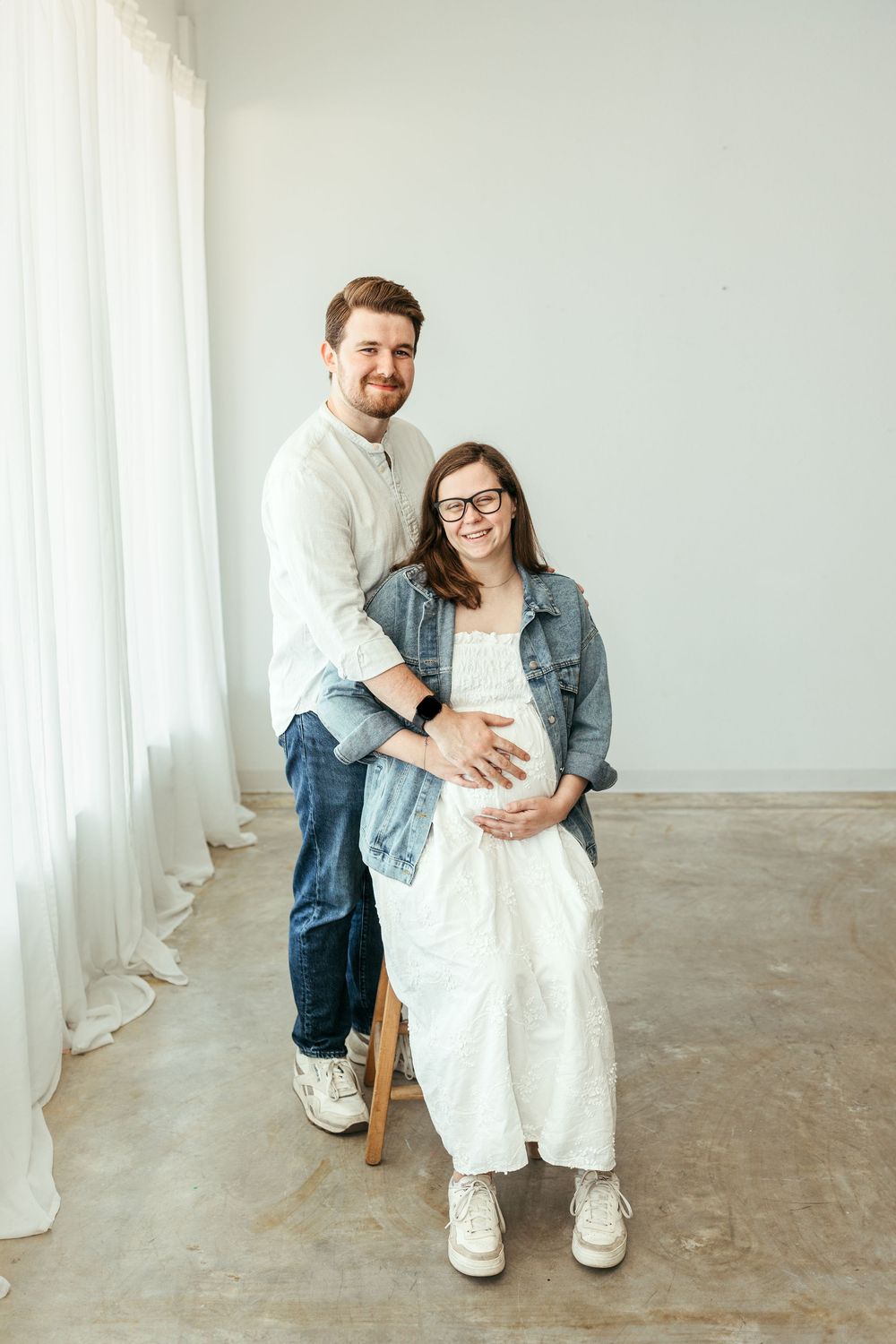 A smiling couple poses in a bright studio, the pregnant woman seated as her partner stands behind her, both cradling her baby bump.