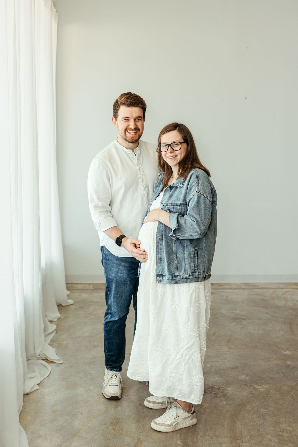 Smiling couple posing together, pregnant woman in white dress and denim jacket, man in white shirt and jeans, bright studio.