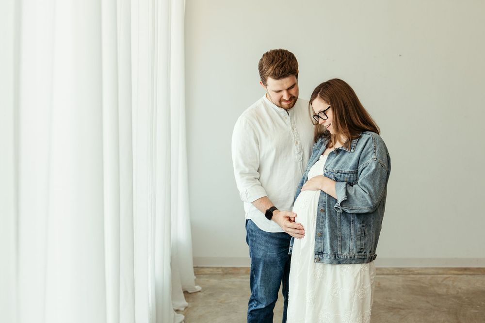 Smiling couple in casual clothes touching pregnant belly in bright white studio room.