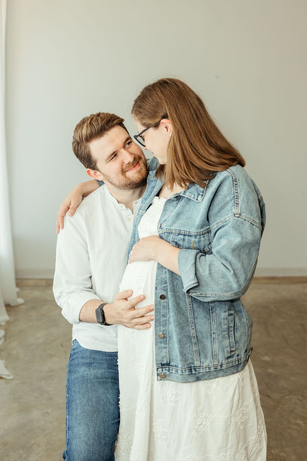 A couple shares a tender moment, with a pregnant woman in a denim jacket embracing her smiling partner indoors.