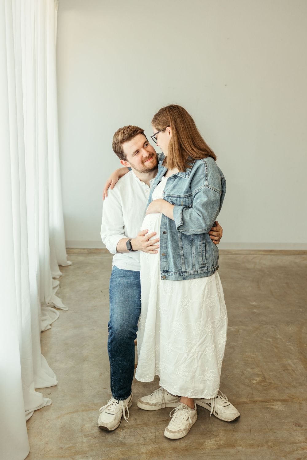 A couple embracing tenderly, pregnant woman in white dress and denim jacket with her partner in white shirt and jeans.